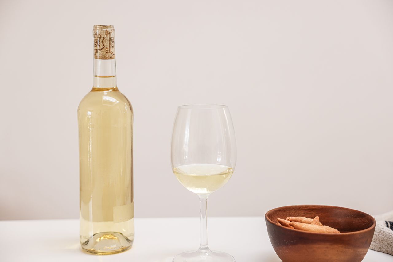 A bottle and glass of white wine alongside a wooden bowl with snacks, set on a minimalistic table.
